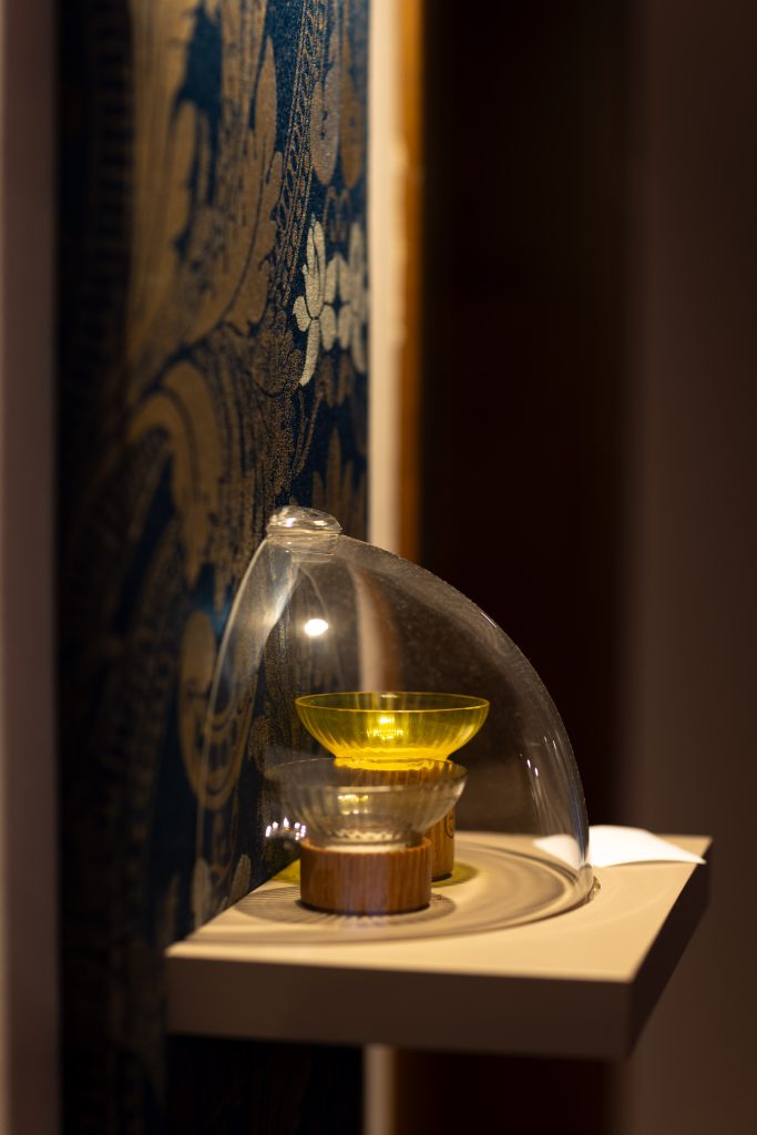 Portacandele esposti Galleria Pieces of Venice – San Servolo Venezia Portacandele di design sostenibile della Galleria Pieces of Venice a San Servolo, esposto sotto cupola in vetro davanti a un tessuto Rubelli